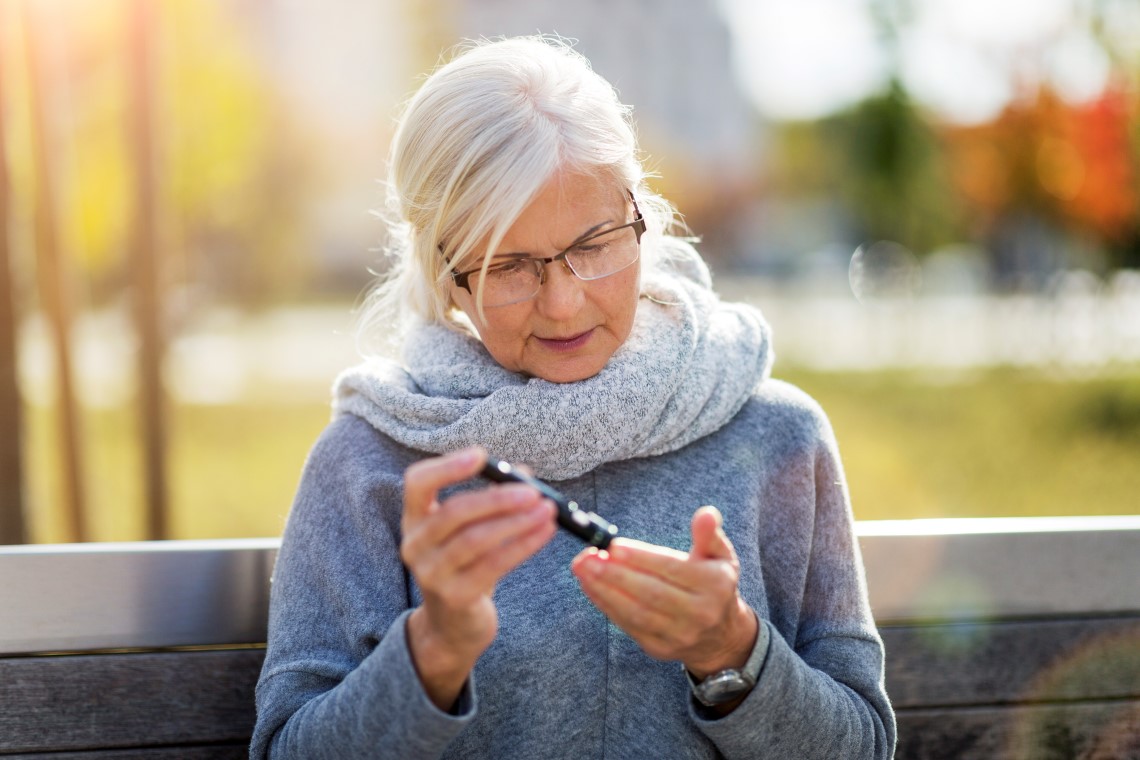 Senior kvinne måler blodsukker ved å stikke seg i fingeren, mens hun sitter på en benk ute.